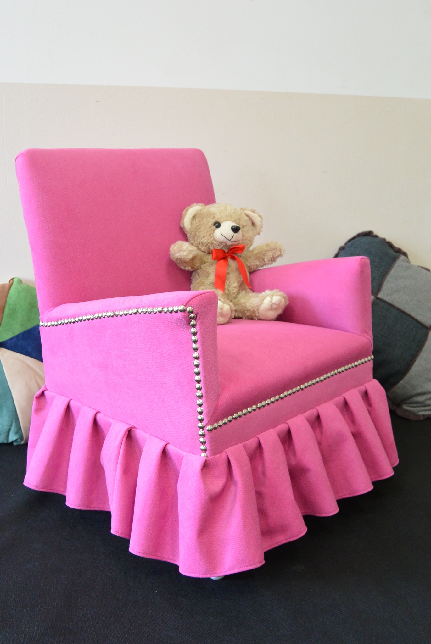 Różowy fotel tapicerowany z falbaną i ozdobnymi ćwiekami, na siedzisku pluszowy miś z czerwoną wstążką. Dekoracyjne poduszki w tle.