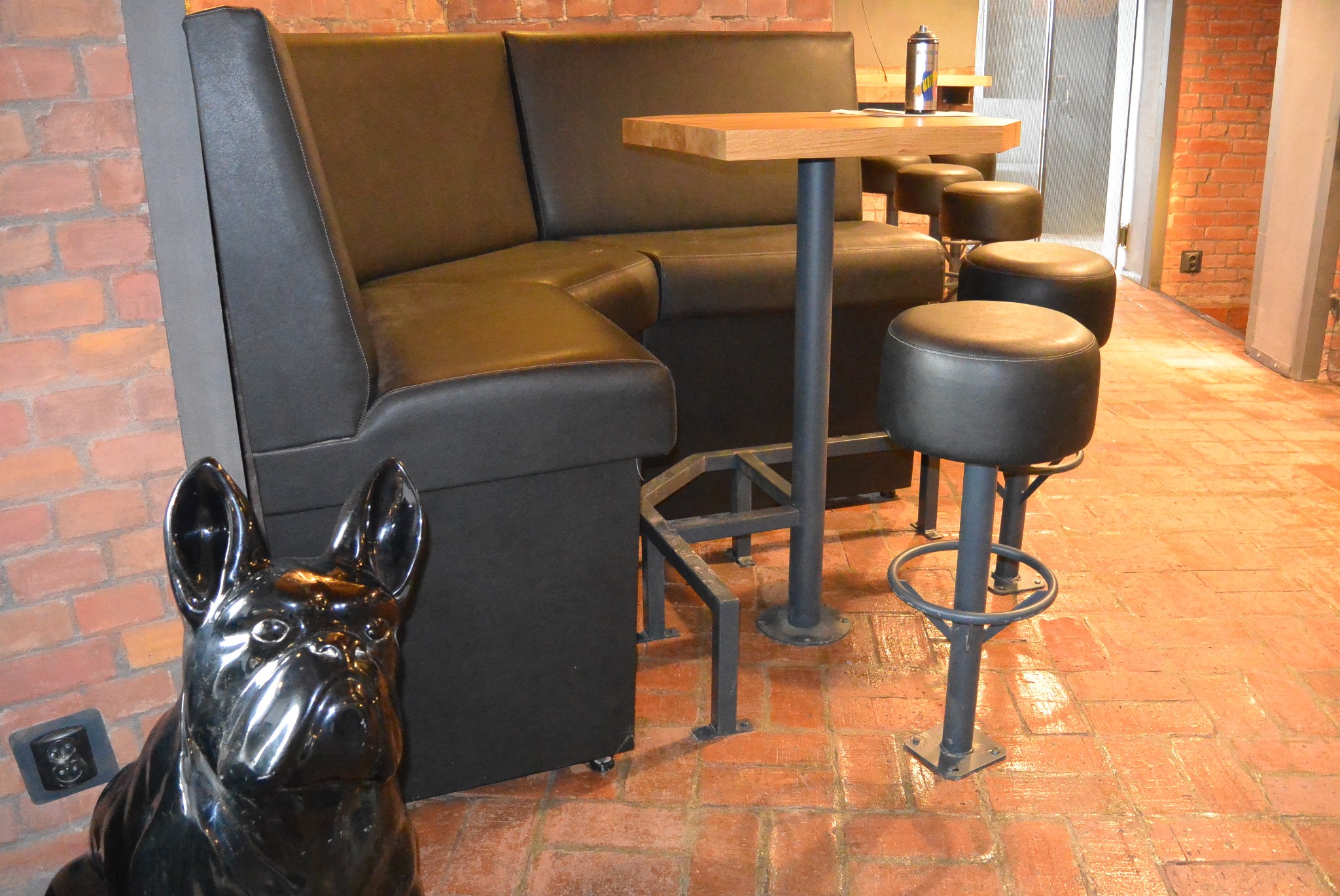 Ciemna, narożna kanapa tapicerowana skórą, stolik barowy i stołki na tle ceglanej ściany. W rogu stoi błyszcząca, czarna figura psa.