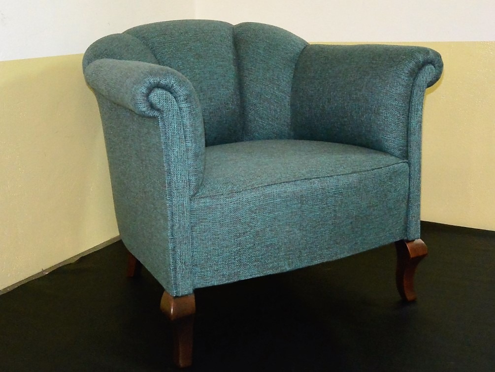 Turkusowy, tapicerowany fotel w stylu retro z drewnianymi nóżkami na ciemnej podłodze, na tle ściany w odcieniach beżu i kremu. Widok z przodu.