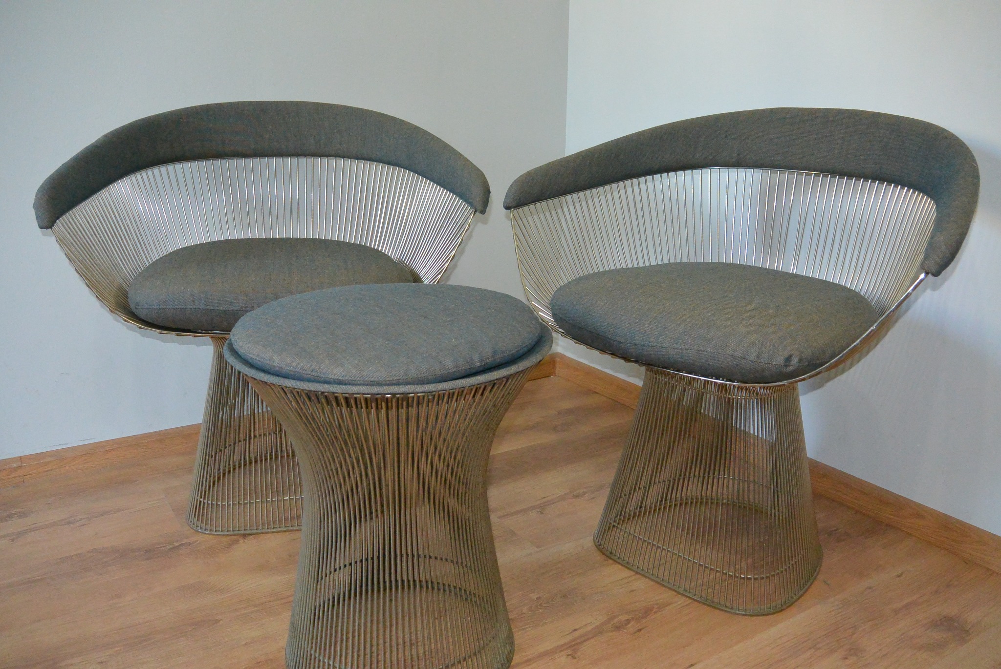 Dwa krzesła i stołek z metalowym szkieletem i szarą tapicerką, ustawione na drewnianej podłodze w rogu pokoju. Minimalistyczny design.