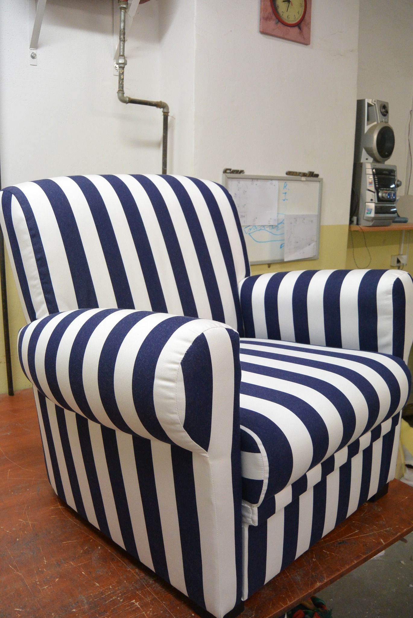 Fotel tapicerowany w biało-granatowe pasy, stojący w pracowni, z widoczną instalacją i sprzętem audio w tle. Ujęcie z niskiego kąta, podkreślające formę mebla.