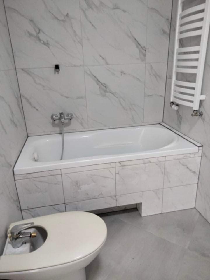 Wykończona łazienka w bieli i szarości: wanna obudowana płytkami imitującymi marmur, grzejnik drabinkowy i bidet. Minimalistyczny styl, świeżo po remoncie.