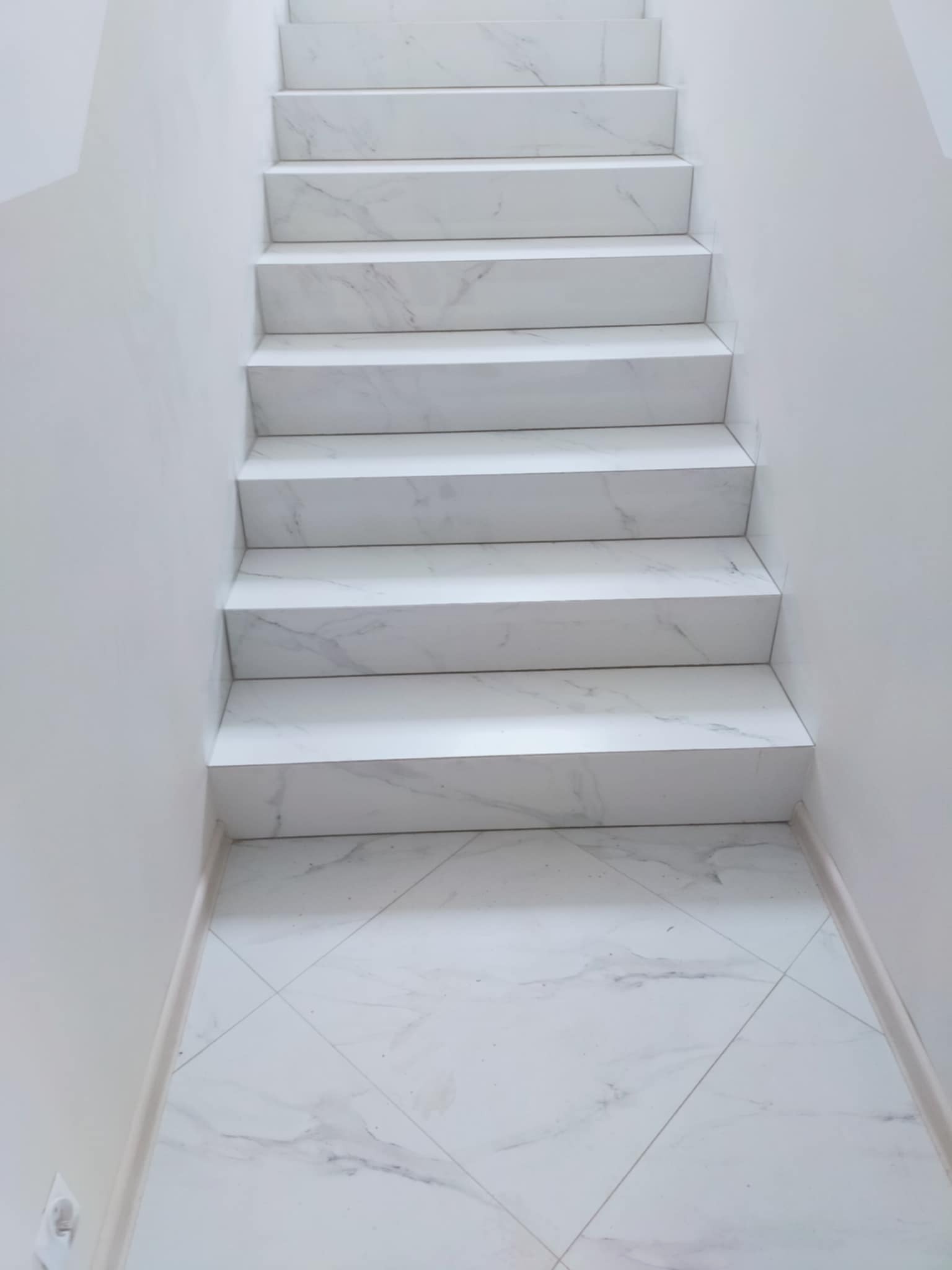 Białe schody z marmurowymi płytkami w wąskim korytarzu, widok z dołu. Płytki podłogowe z wzorem rombów. Minimalistyczny design, jasne wnętrze.