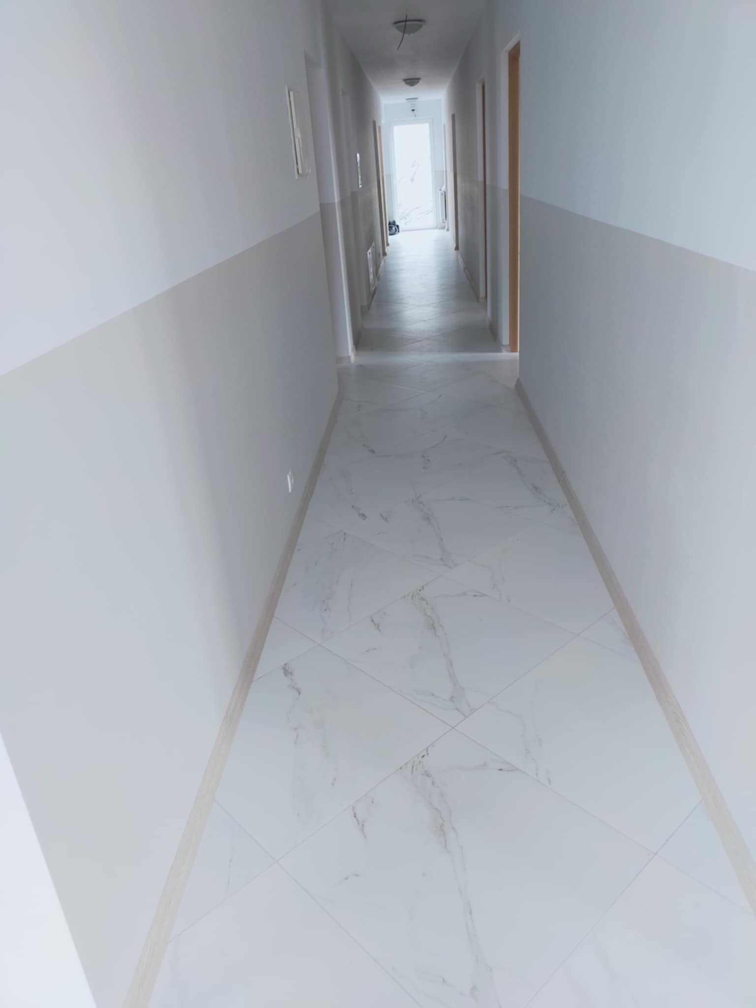 Wykończony korytarz z białymi płytkami imitującymi marmur i dwukolorowymi ścianami, jasne wnętrze z widocznymi drzwiami i oświetleniem sufitowym w głębi.