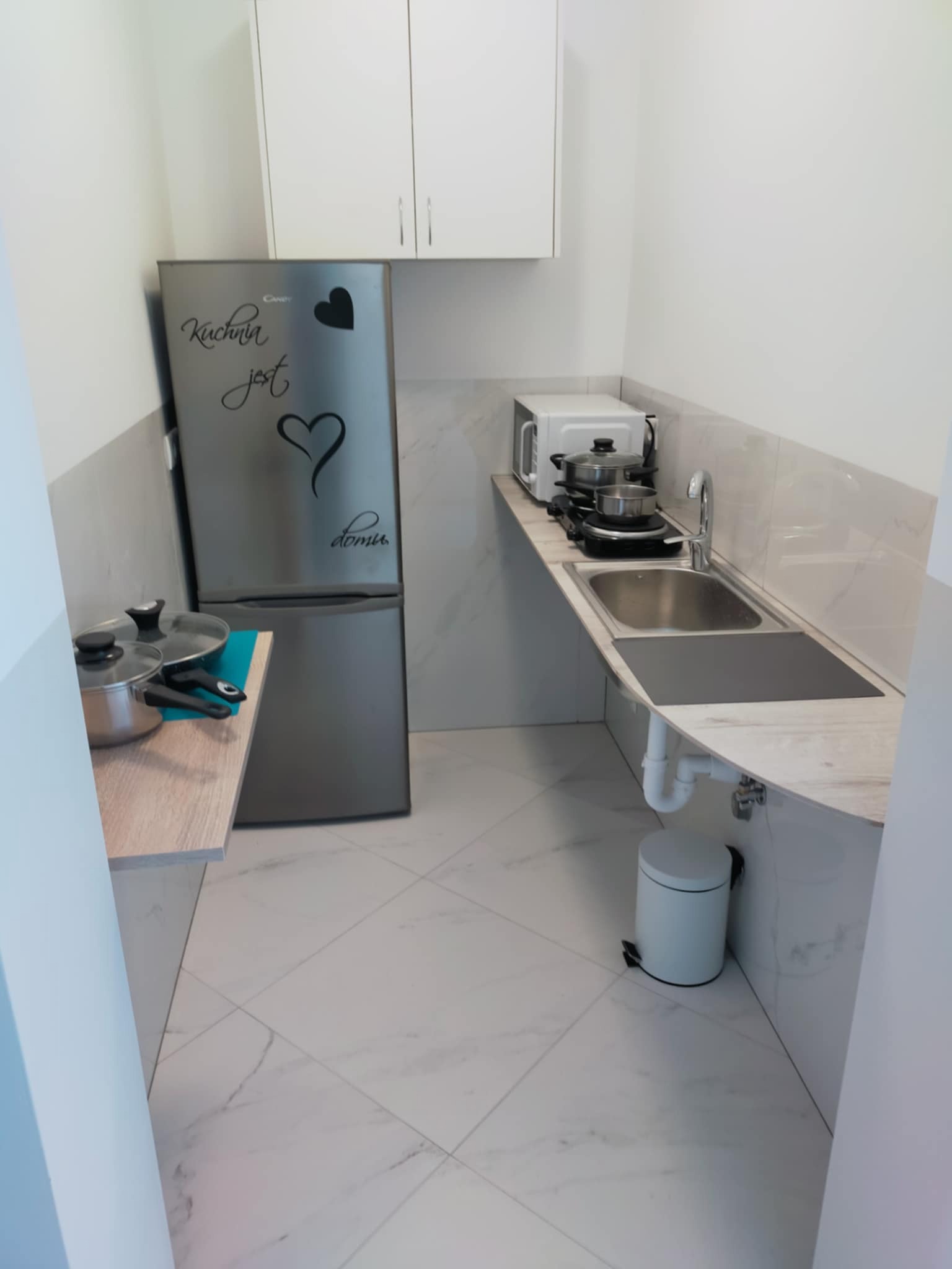 Jasna kuchnia z marmurową podłogą i blatem. Na blacie zlewozmywak, płyta indukcyjna, mikrofalówka i garnki. Na lodówce naklejki. Białe szafki wiszące.