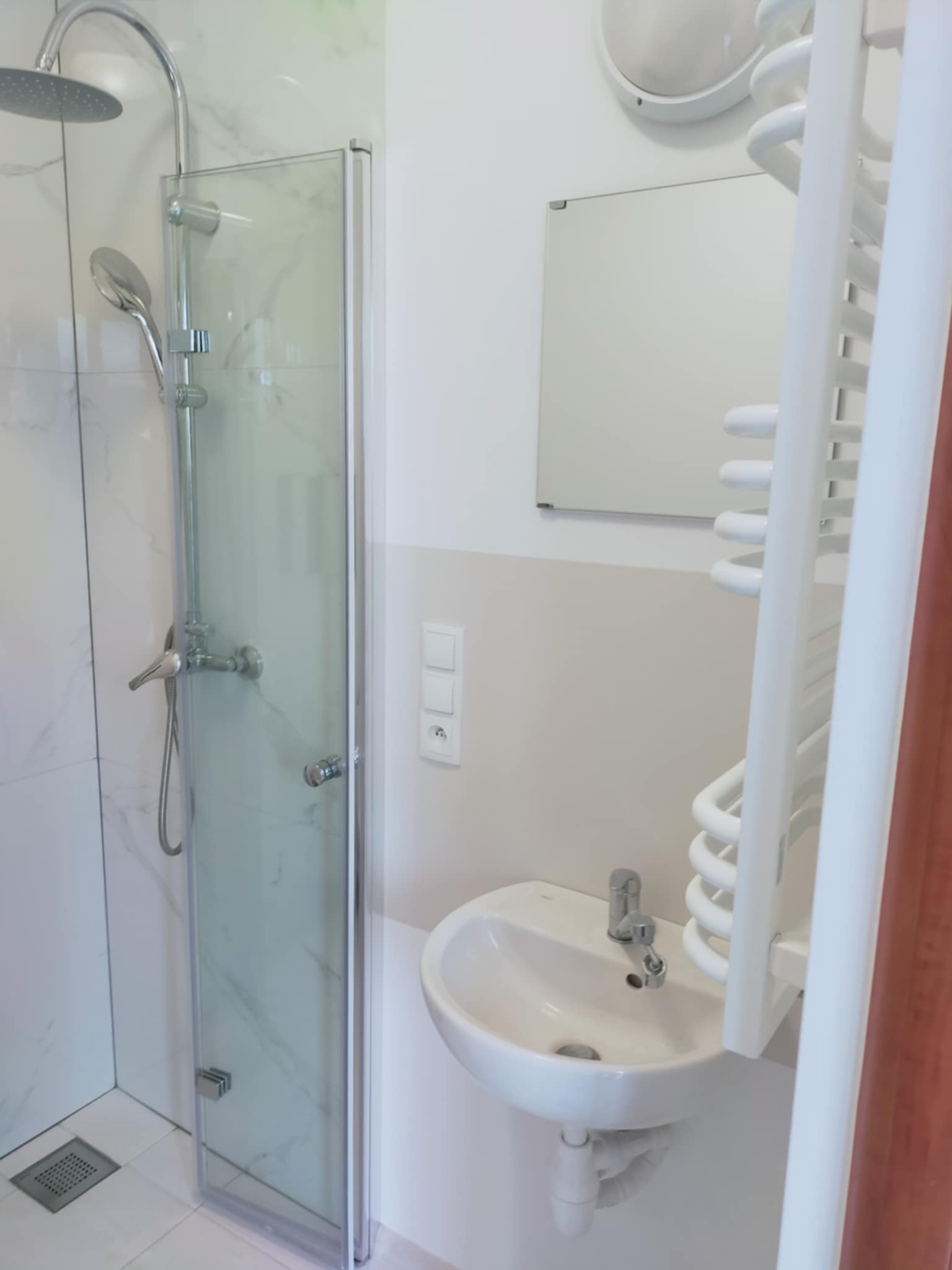 Nowoczesna łazienka z kabiną prysznicową typu walk-in, białą umywalką i grzejnikiem drabinkowym. Minimalistyczny design, marmurowe płytki.
