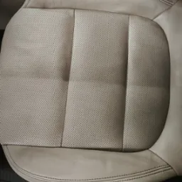 Beżowe, perforowane skórzane siedzenie samochodowe z wyraźnymi szwami, ukazane z góry. Widoczny fragment dolnej krawędzi siedziska i fragment podłogi pojazdu.