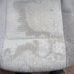 Janek Nowrocki - Szare, mocno zabrudzone siedzisko samochodowe z widocznymi plamami, sfotografowane z góry. W dolnej części kadru widoczne buty osoby stojącej przed fotelem.