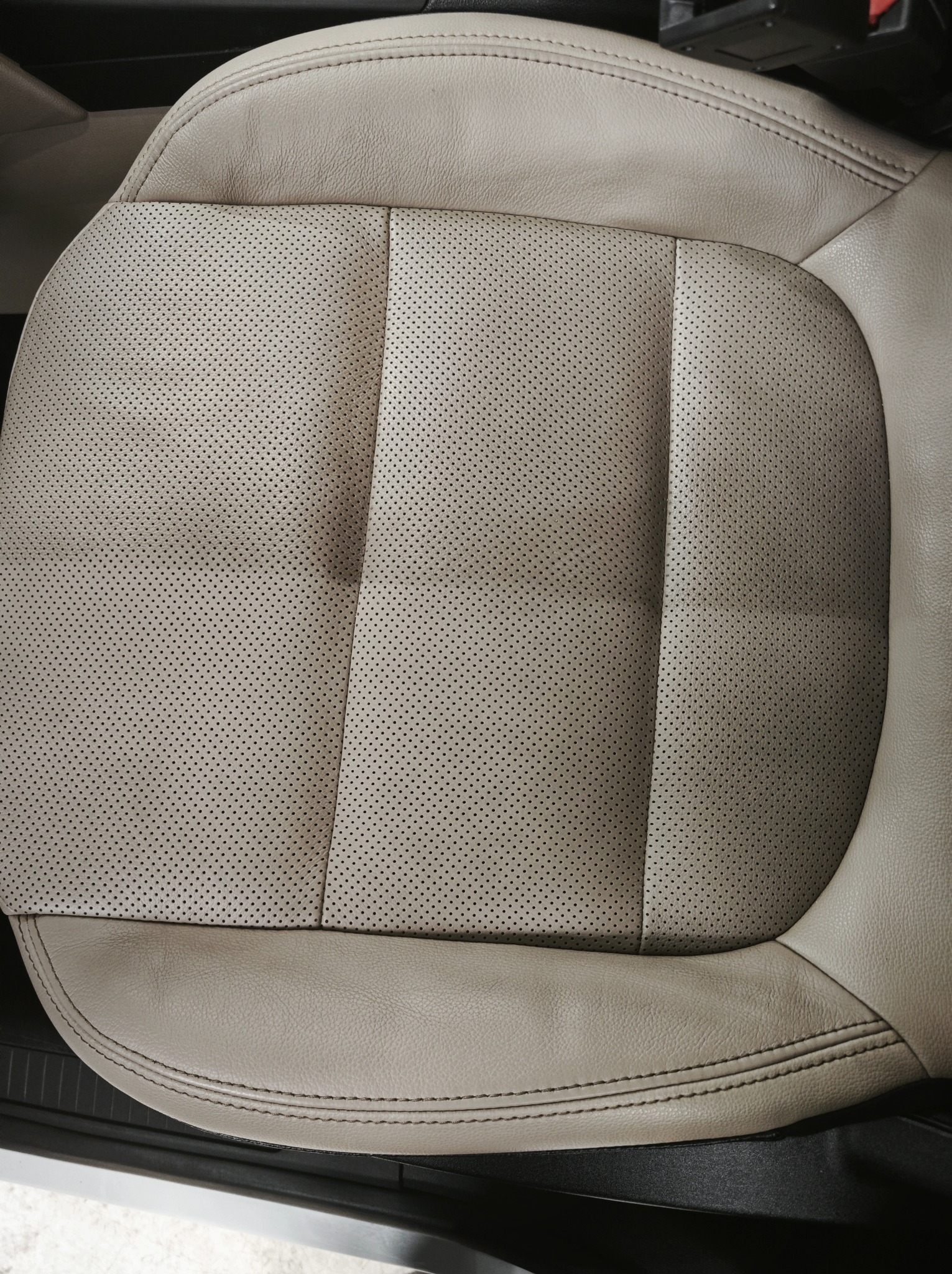Beżowe, perforowane skórzane siedzenie samochodowe z wyraźnymi szwami, ukazane z góry. Widoczny fragment dolnej krawędzi siedziska i fragment podłogi pojazdu.