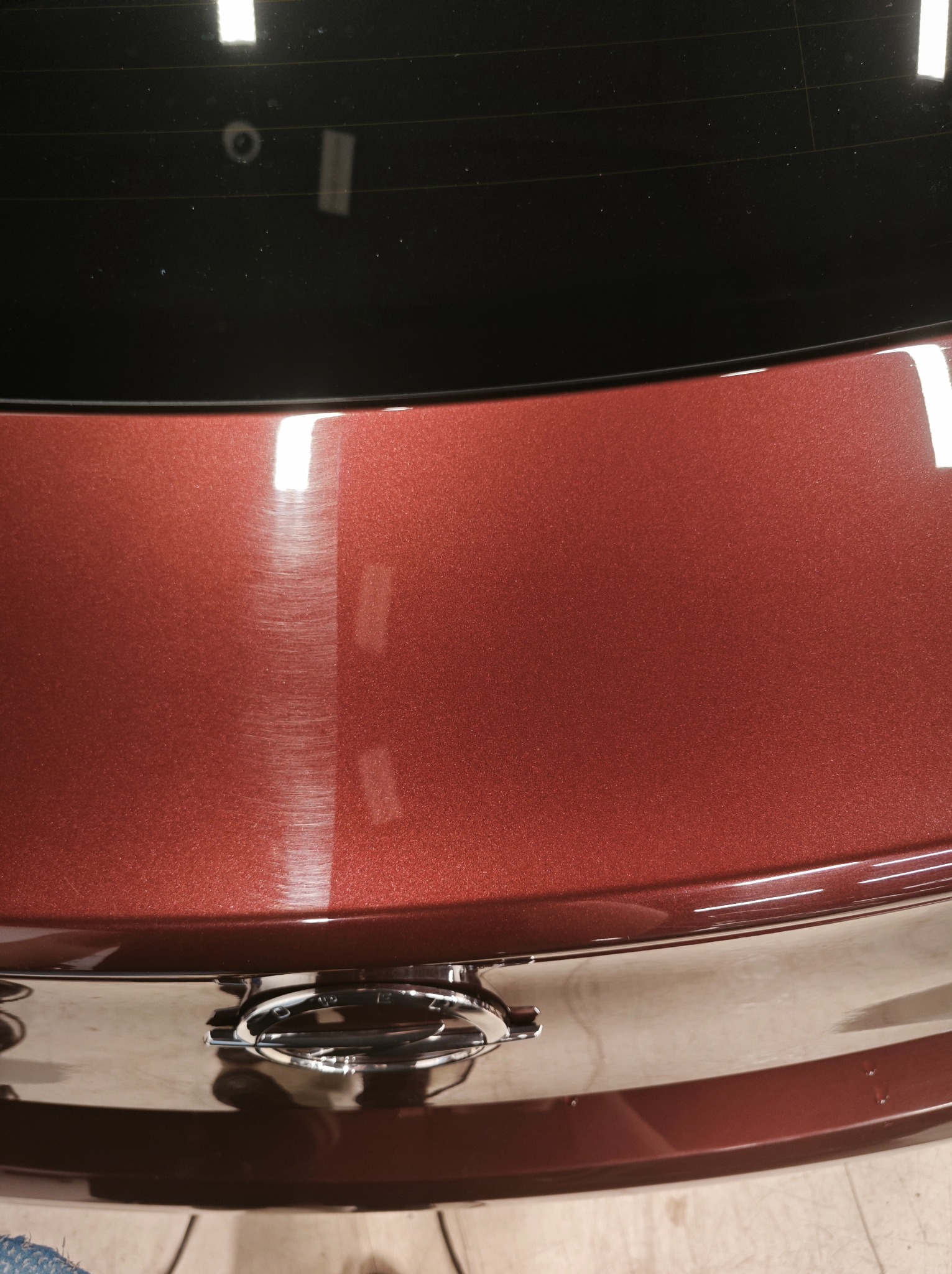 Błyszcząca, bordowa karoseria samochodu po polerowaniu, widoczne odbicia światła, chromowany zamek bagażnika, fragment szyby i podłogi garażu.