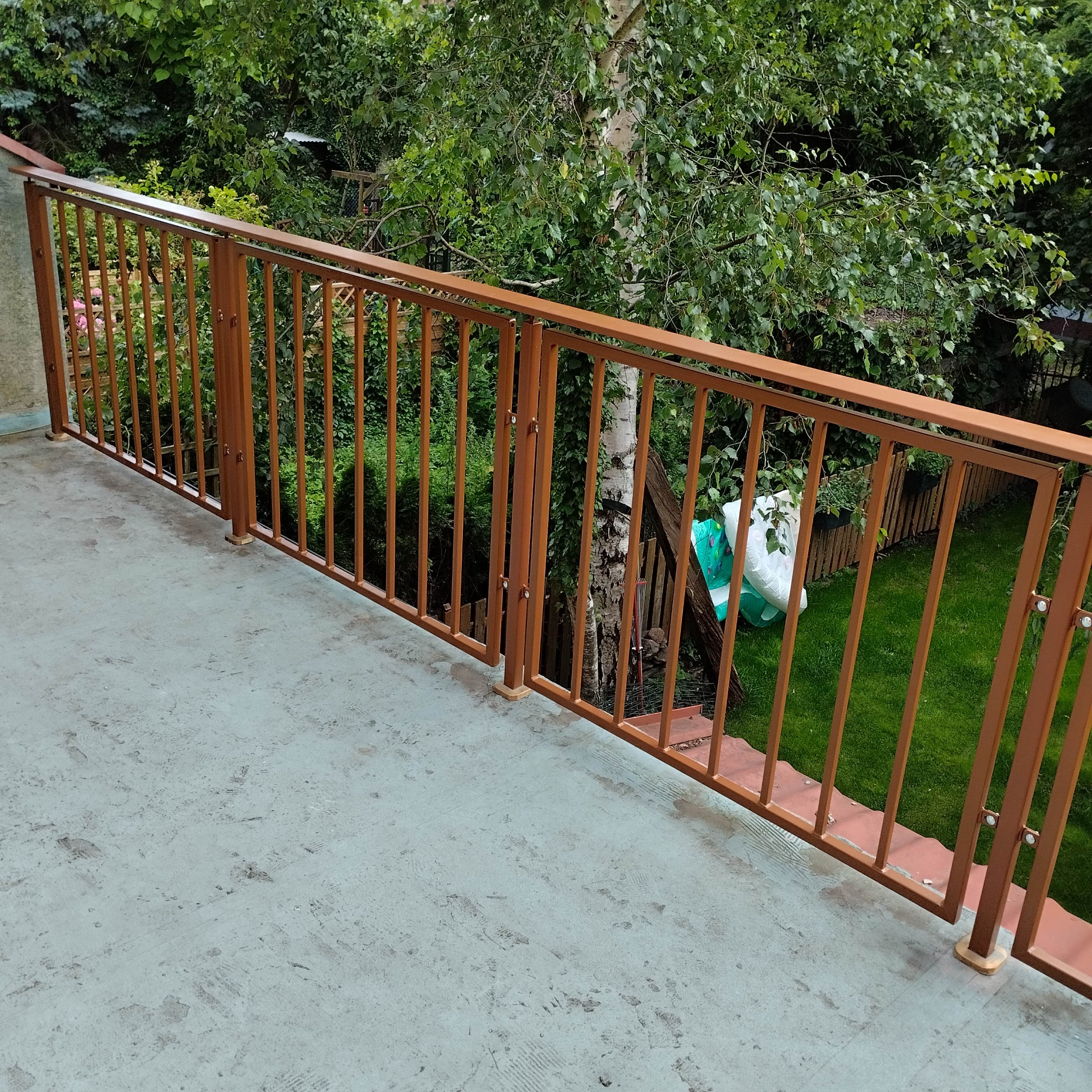 Balkon z brązową, metalową balustradą z pionowymi szczebelkami. Za balustradą widoczny ogród z drzewami i trawą. Ujęcie z góry, w dziennym świetle.