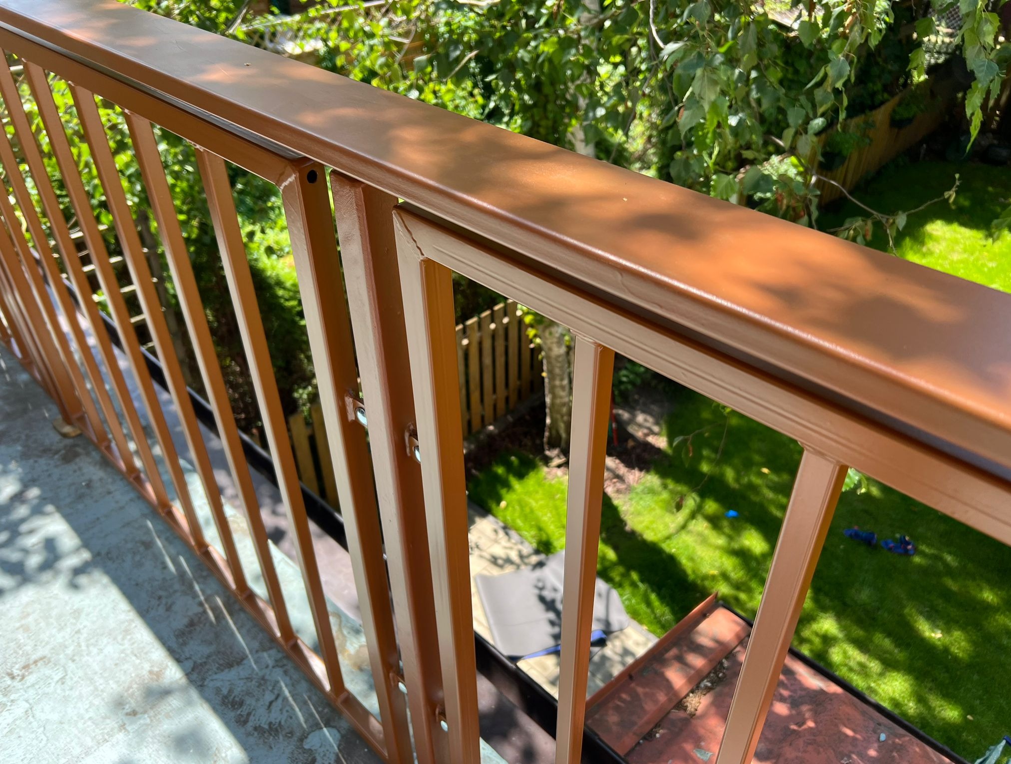 Balkon z brązową, metalową balustradą, widok z góry na zielony ogród. Elementy mocujące widoczne na balustradzie.