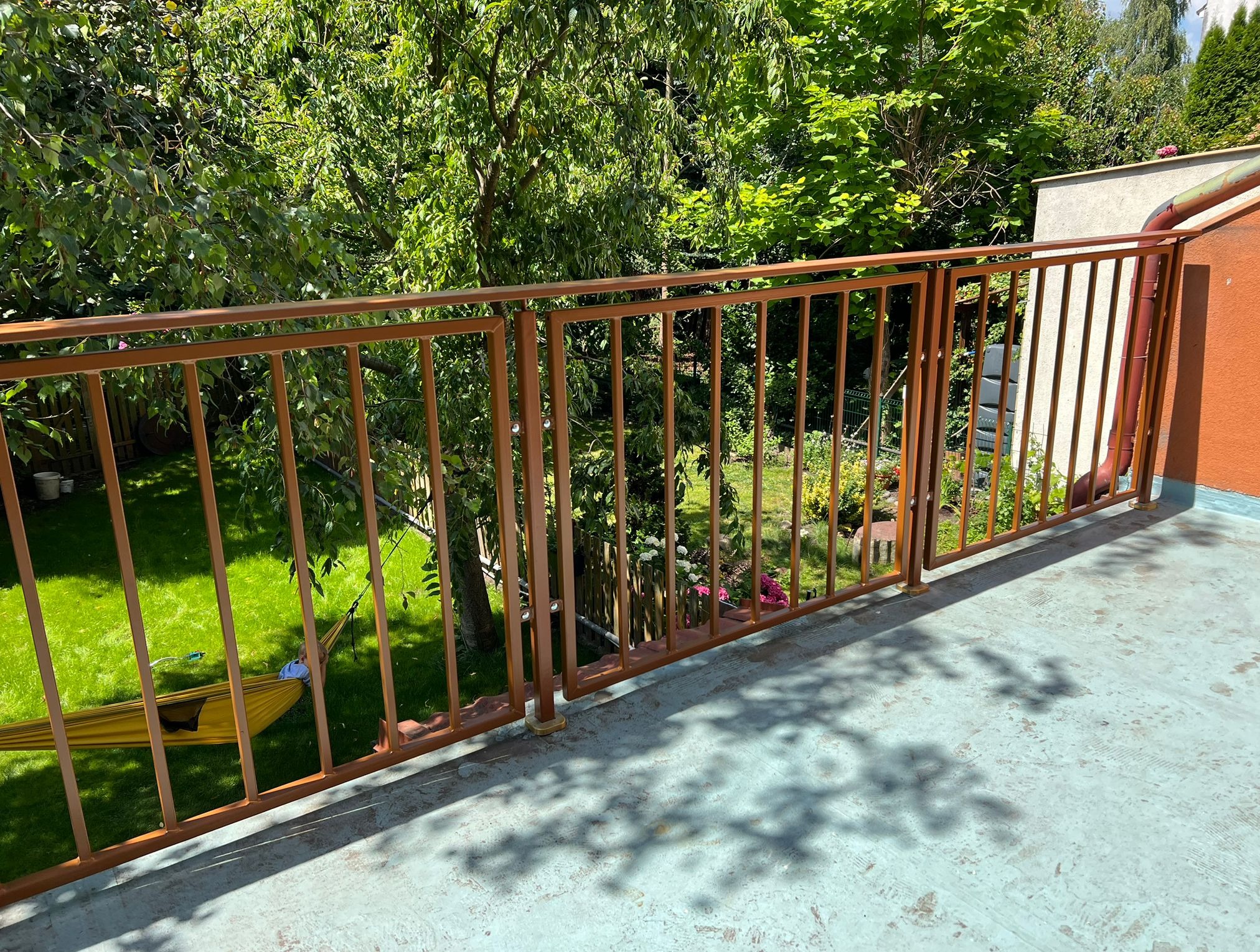 Balkon z metalową, brązową balustradą na tle zielonego ogrodu. W oddali widoczny hamak i drzewa. Ujęcie z góry, oświetlone naturalnym światłem.