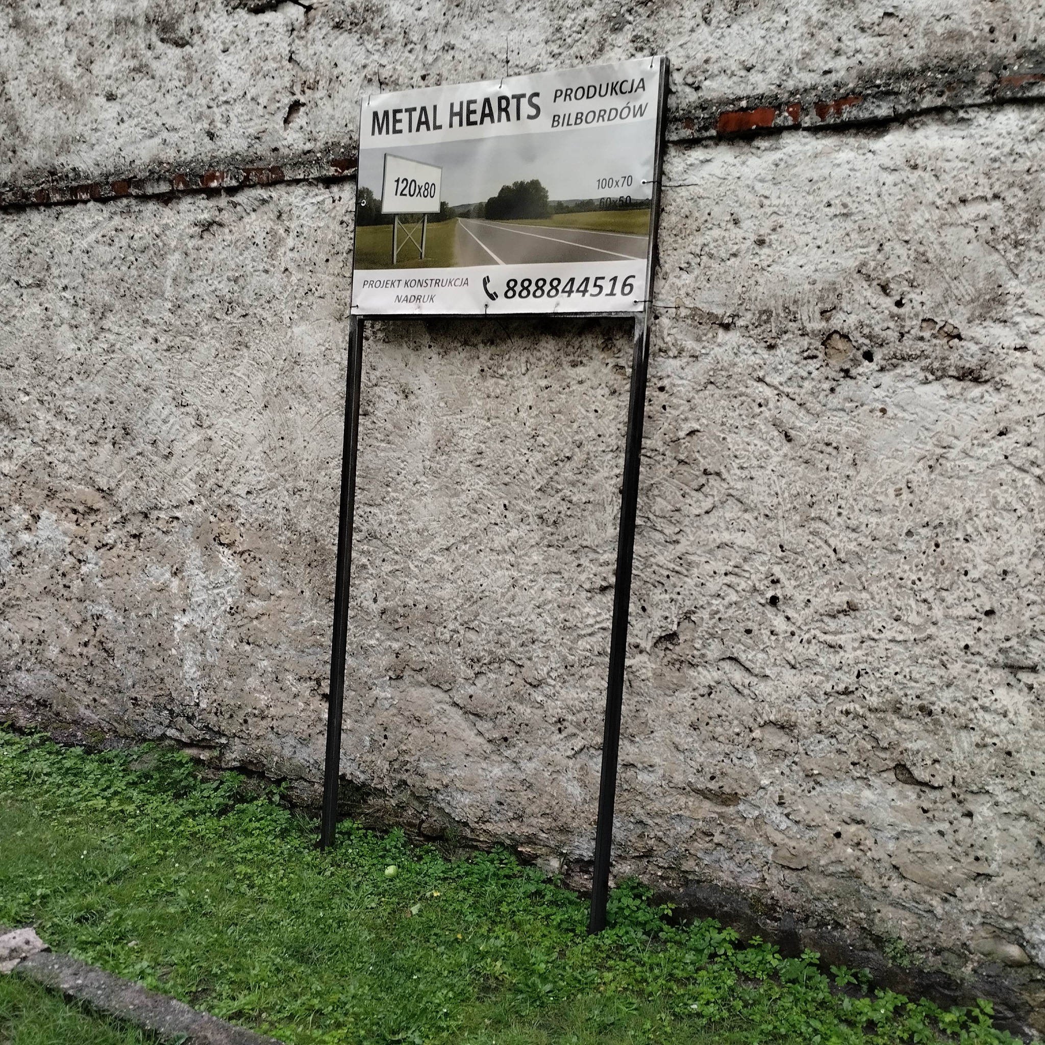 Metalowy stelaż z tablicą reklamową producenta bilbordów na tle szarego, chropowatego muru i trawy. Na tablicy widnieją wymiary i zdjęcie drogi.