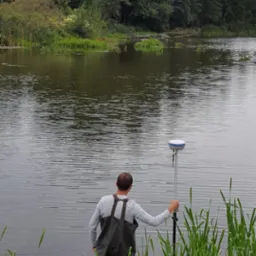 Pracownik w wodoodpornych spodniobutach, trzymający tyczkę z odbiornikiem GPS na tle jeziora i zieleni, wykonuje pomiary geodezyjne. Odbiornik GPS.