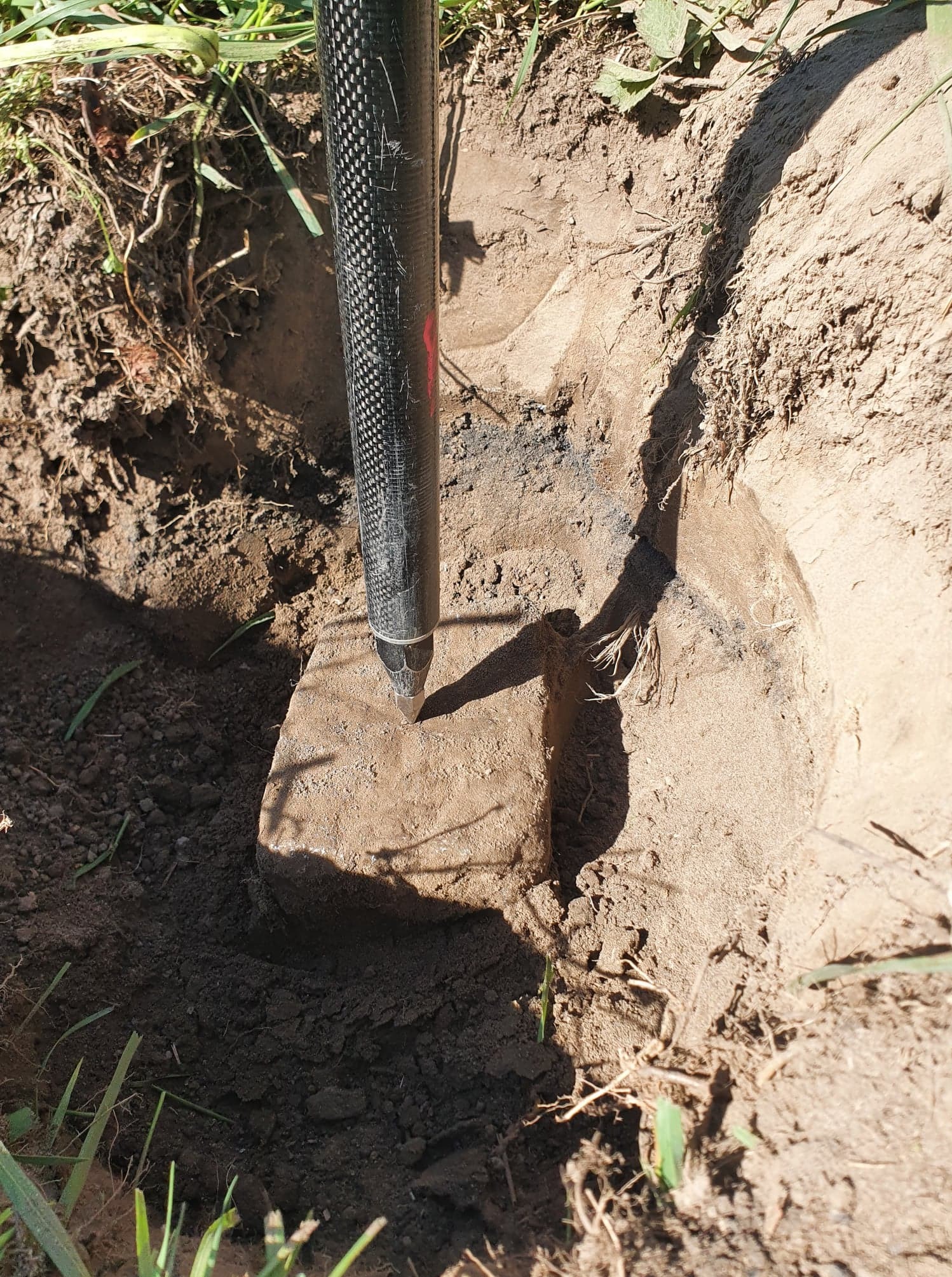 Wbity w ziemię czarny tyczkowy reper z czerwonym oznaczeniem, używany do precyzyjnych pomiarów wysokościowych w geodezji. Widoczny otwór w ziemi.