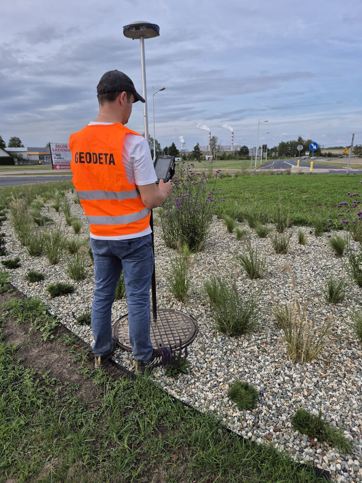 Pomiar geodezyjny na rondzie: mężczyzna w kamizelce z napisem 'Geodeta' obsługuje sprzęt pomiarowy na tle zieleni i infrastruktury drogowej.