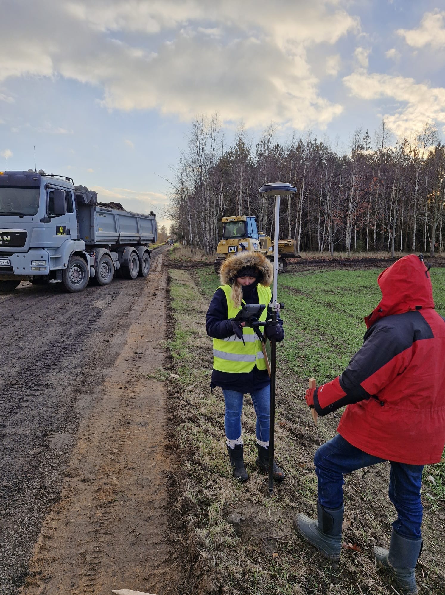 Praca w terenie: kobieta w kamizelce mierzy dane za pomocą precyzyjnego instrumentu pomiarowego, w tle ciężarówka MAN i koparka CAT.