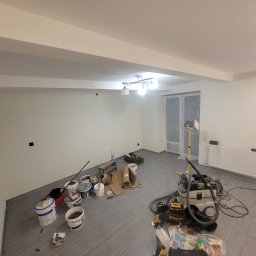 Uslugi remontowe Jakub Misior - Wnętrze w trakcie malowania ścian na biało. Widoczne narzędzia malarskie, drabina, grzejnik i osłonięte folią drzwi balkonowe. Szare panele na podłodze i lampa sufitowa.