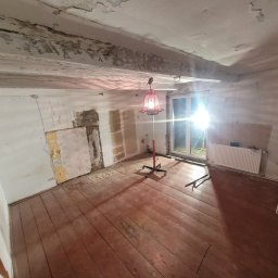Uslugi remontowe Jakub Misior - Wnętrze pokoju w trakcie remontu: odsłonięte ściany z fragmentami tapet, drewniana podłoga, grzejnik i drabina oparta o ścianę. Centralnie stoi lampa podłogowa.