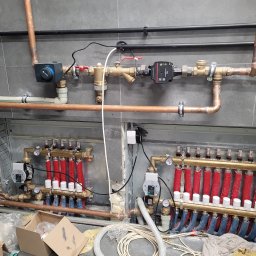 Uslugi remontowe Jakub Misior - Rozdzielacz ogrzewania podłogowego z rurami w izolacji czerwonej i niebieskiej, widoczne zawory i manometry, instalacja w szarej płytce.