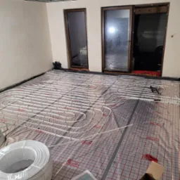 Białe rury ogrzewania podłogowego Roth ułożone na folii z siatką w surowym wnętrzu. Widoczne zwoje rur i narzędzia. Przez okno widać dostawczy samochód.