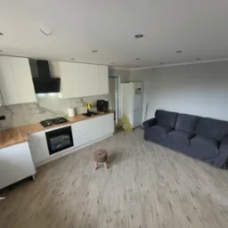 Jasne, przestronne wnętrze po remoncie: kuchnia z białymi szafkami, drewnianym blatem i marmurową ścianą, połączona z salonem z szarą sofą i drewnianą podłogą.