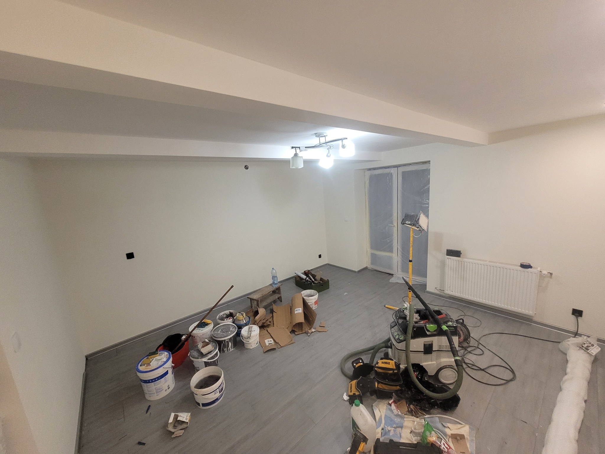 Wnętrze w trakcie malowania ścian na biało. Widoczne narzędzia malarskie, drabina, grzejnik i osłonięte folią drzwi balkonowe. Szare panele na podłodze i lampa sufitowa.