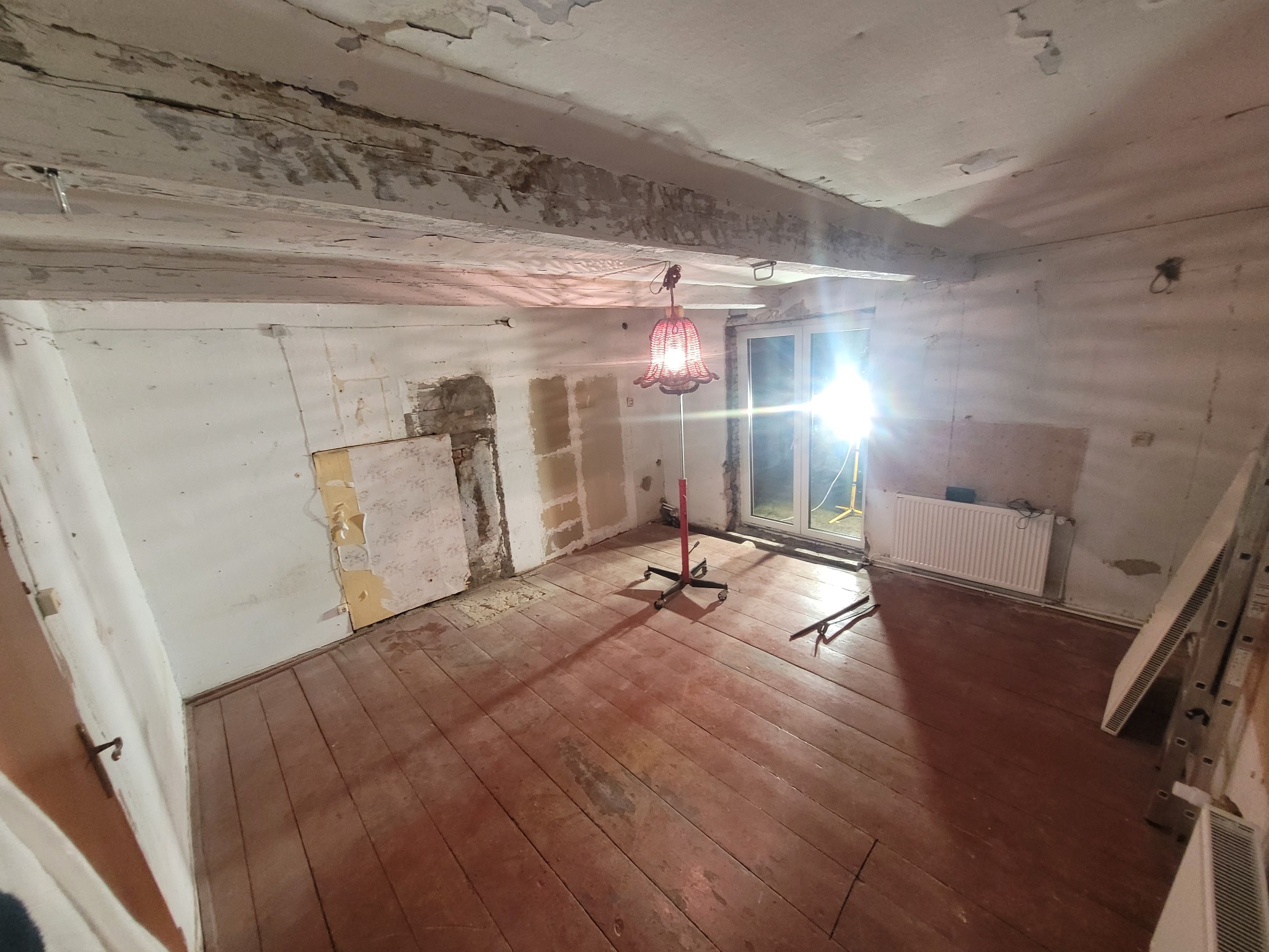 Wnętrze pokoju w trakcie remontu: odsłonięte ściany z fragmentami tapet, drewniana podłoga, grzejnik i drabina oparta o ścianę. Centralnie stoi lampa podłogowa.