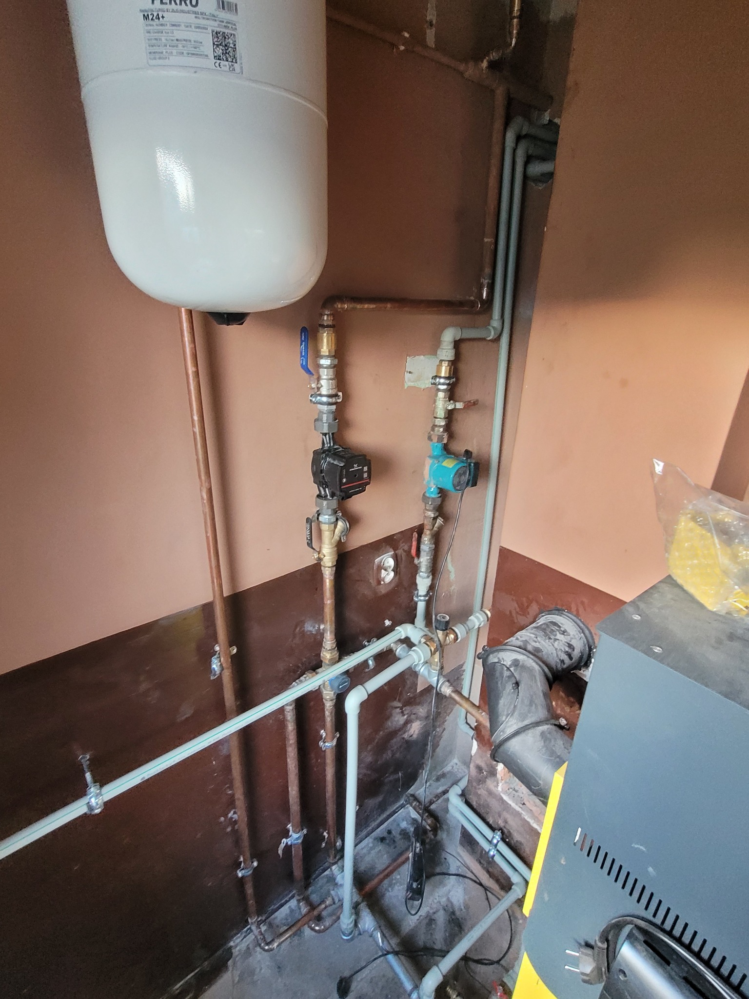 Instalacja grzewcza z białym zasobnikiem FERRO, rurami miedzianymi i plastikowymi. Widoczne pompy obiegowe i fragment kotła. Ujęcie z dołu, przy ścianie w brązowym odcieniu.