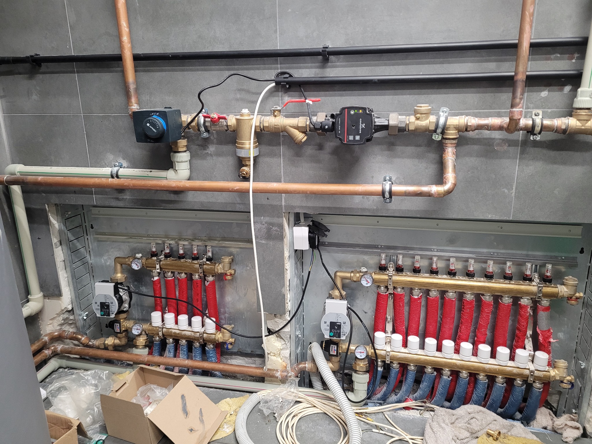 Rozdzielacz ogrzewania podłogowego z rurami w izolacji czerwonej i niebieskiej, widoczne zawory i manometry, instalacja w szarej płytce.