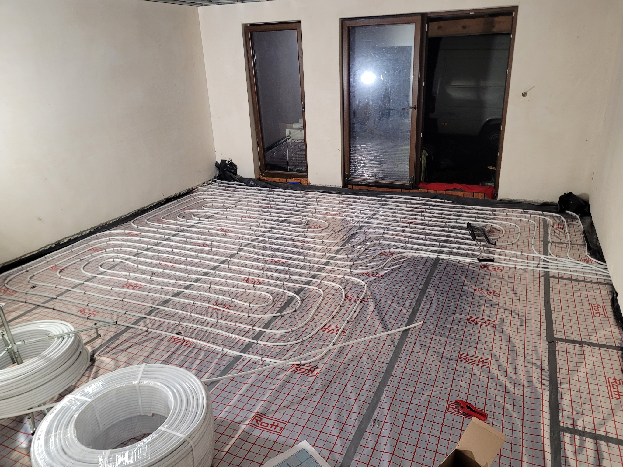 Białe rury ogrzewania podłogowego Roth ułożone na folii z siatką w surowym wnętrzu. Widoczne zwoje rur i narzędzia. Przez okno widać dostawczy samochód.