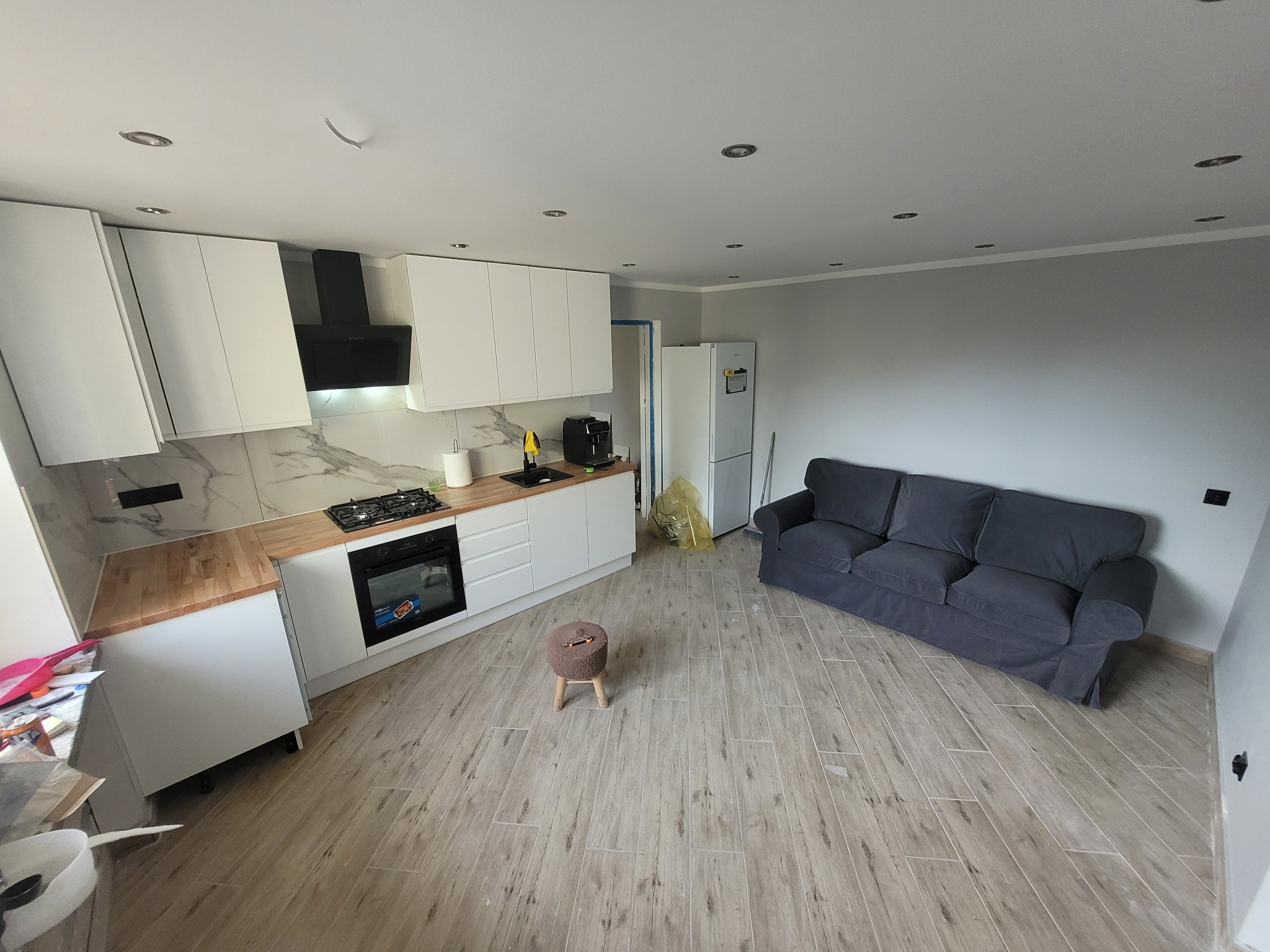 Jasne, przestronne wnętrze po remoncie: kuchnia z białymi szafkami, drewnianym blatem i marmurową ścianą, połączona z salonem z szarą sofą i drewnianą podłogą.