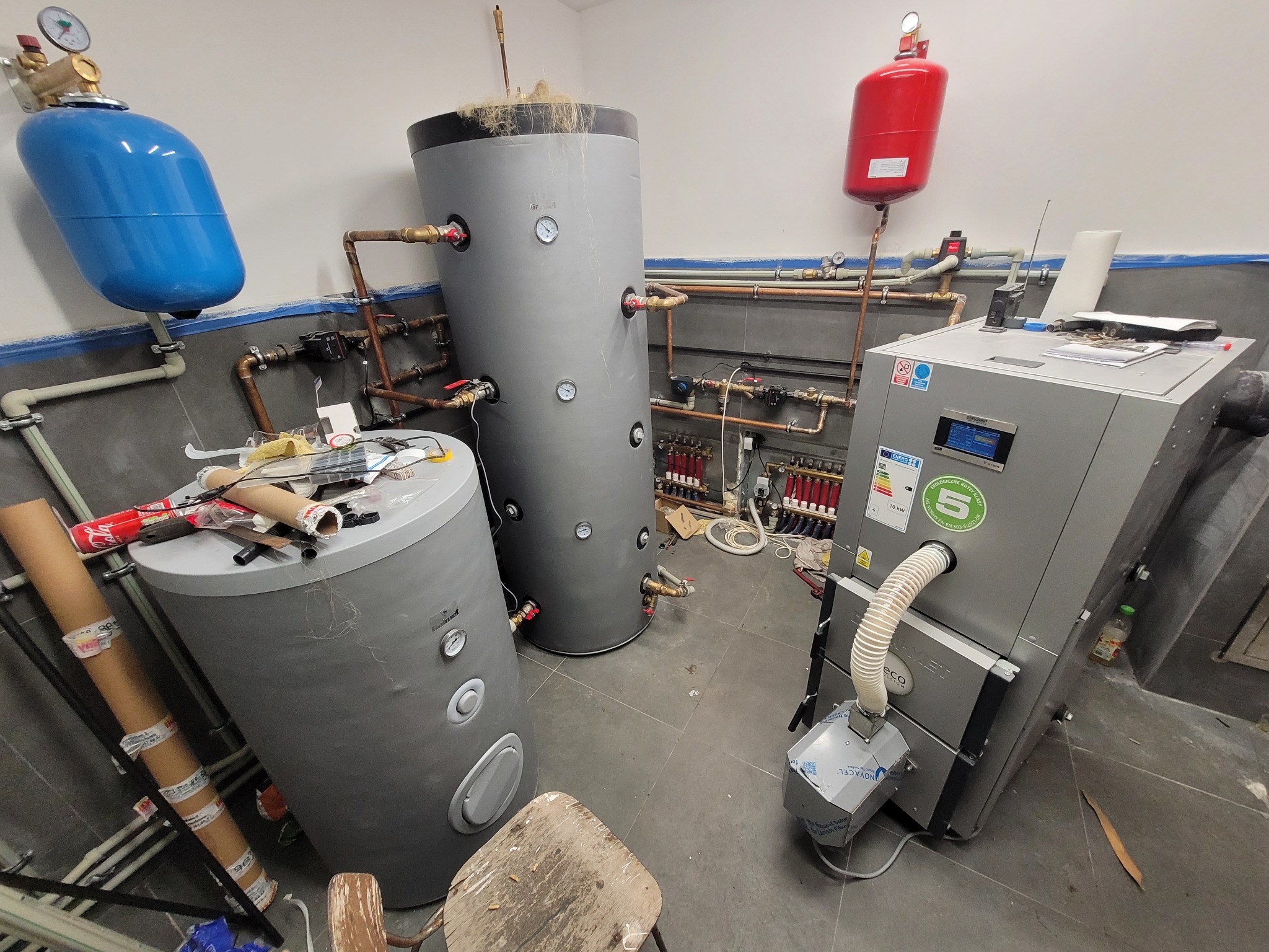 Instalacja kotła CO i zasobników wody w kotłowni. Widoczne elementy instalacji grzewczej, rury miedziane, zbiorniki, nowy piec. Na zbiornikach widoczne manometry i zawory.