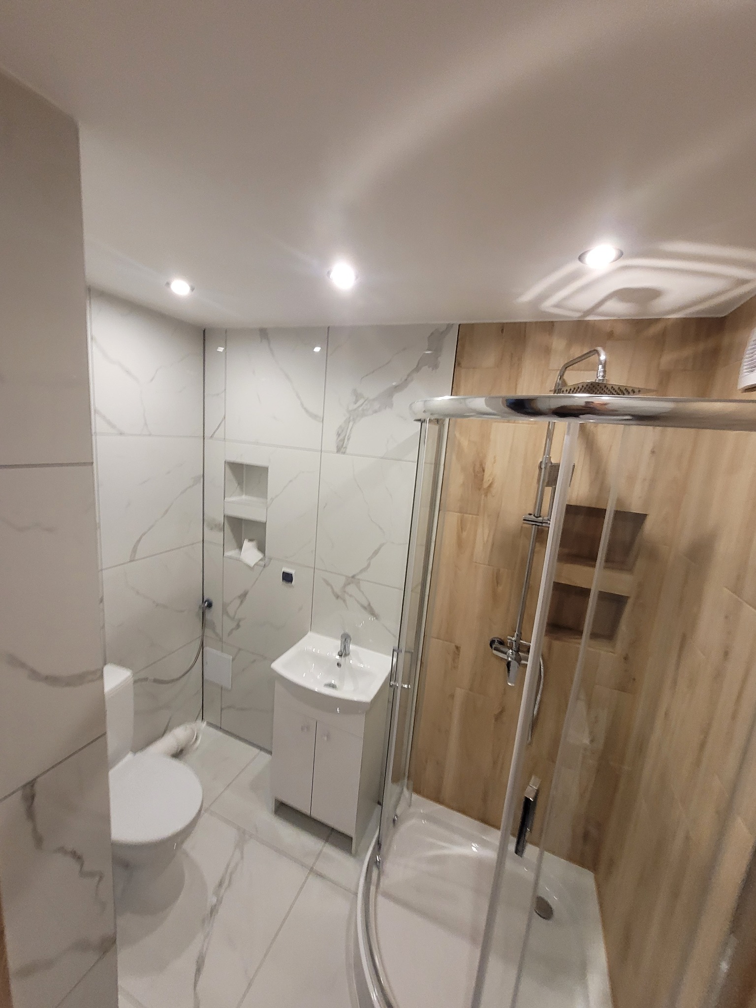 Nowoczesna, jasna łazienka po remoncie: białe marmurowe płytki, kabina prysznicowa ze szklanymi drzwiami, biała szafka z umywalką i toaleta. Oświetlenie punktowe.