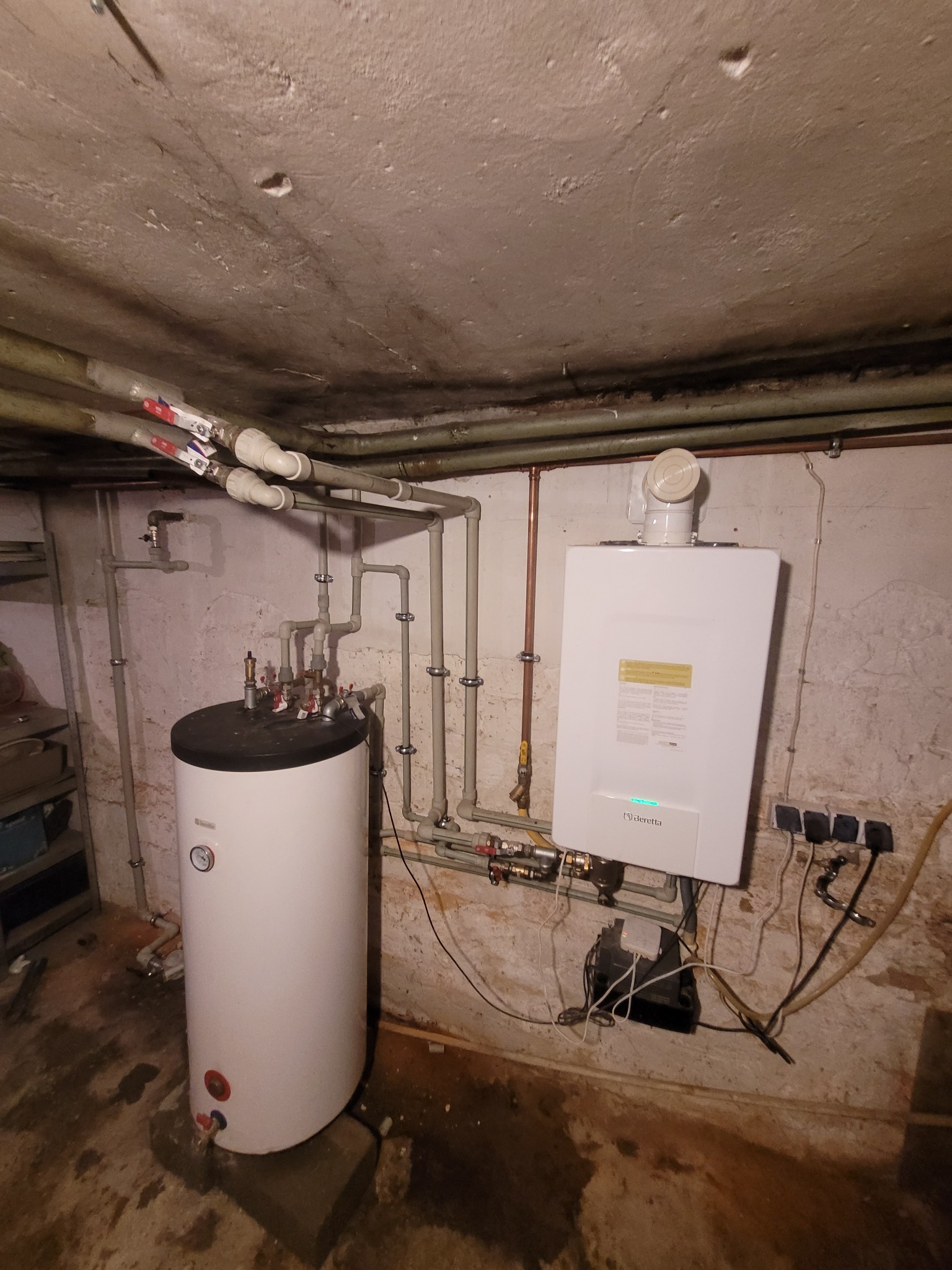 Biały piec grzewczy Beretta i zasobnik ciepłej wody w piwnicy. Widoczne rury instalacji grzewczej i elektryczne. Surowy beton na ścianach i suficie.