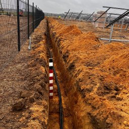 Maobra - Długi wykop w ziemi z czarnymi kablami ułożonymi wzdłuż. W tle konstrukcja pod panele słoneczne i ogrodzenie z siatki. Widoczny znacznik głębokości wykopu.