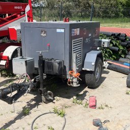 Maobra - Przenośny kompresor budowlany na przyczepie, obok czerwony podnośnik i rury drenarskie. Widoczne narzędzia i wózek transportowy. Sprzęt na placu budowy.