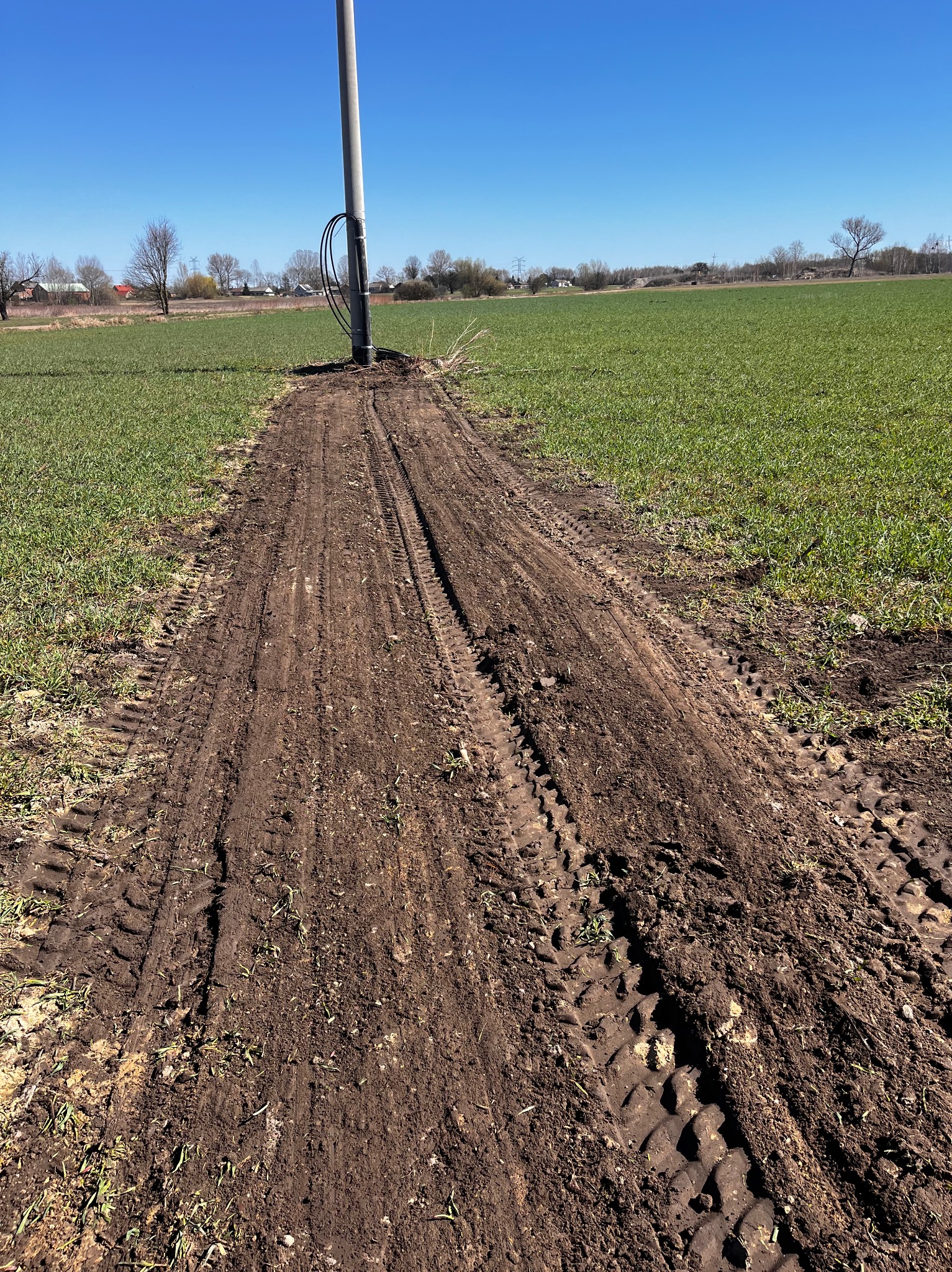 Ślady opon w ziemi prowadzące do słupa energetycznego na tle zielonego pola i błękitnego nieba. Widoczne świeże naruszenie gruntu.