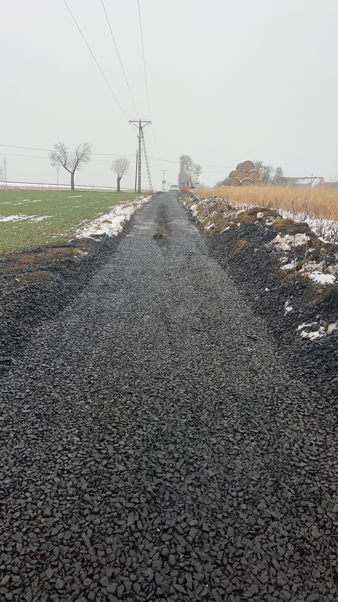 Droga z wysypanym grysem w krajobrazie wiejskim, widoczna koparka w oddali, słupy energetyczne i bele siana. Częściowo zaśnieżony teren, pochmurny dzień.