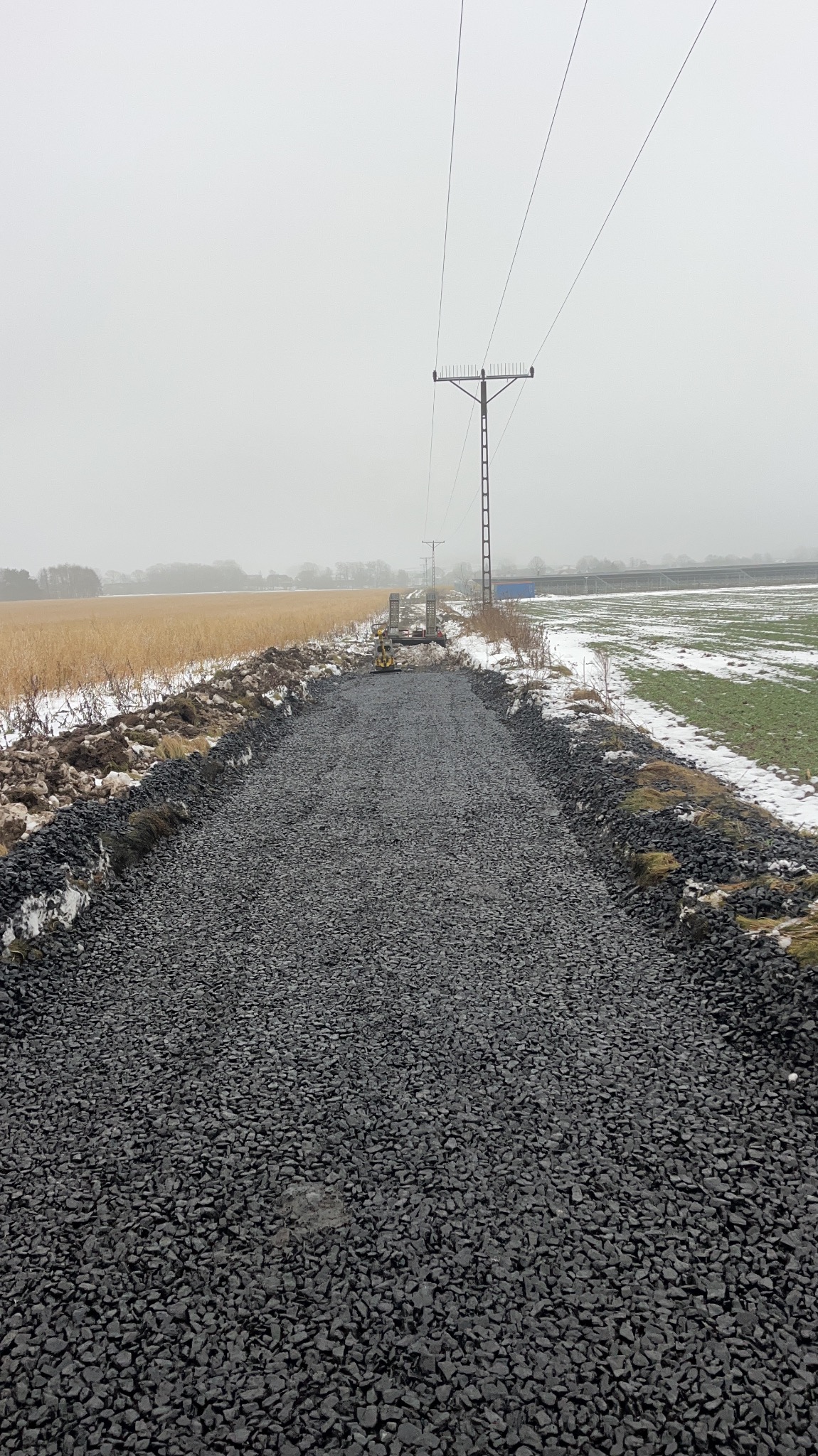 Utwardzona droga z kruszywa w krajobrazie zimowym, widok z perspektywy. Słupy energetyczne w tle. Częściowo ośnieżone pola po bokach drogi.