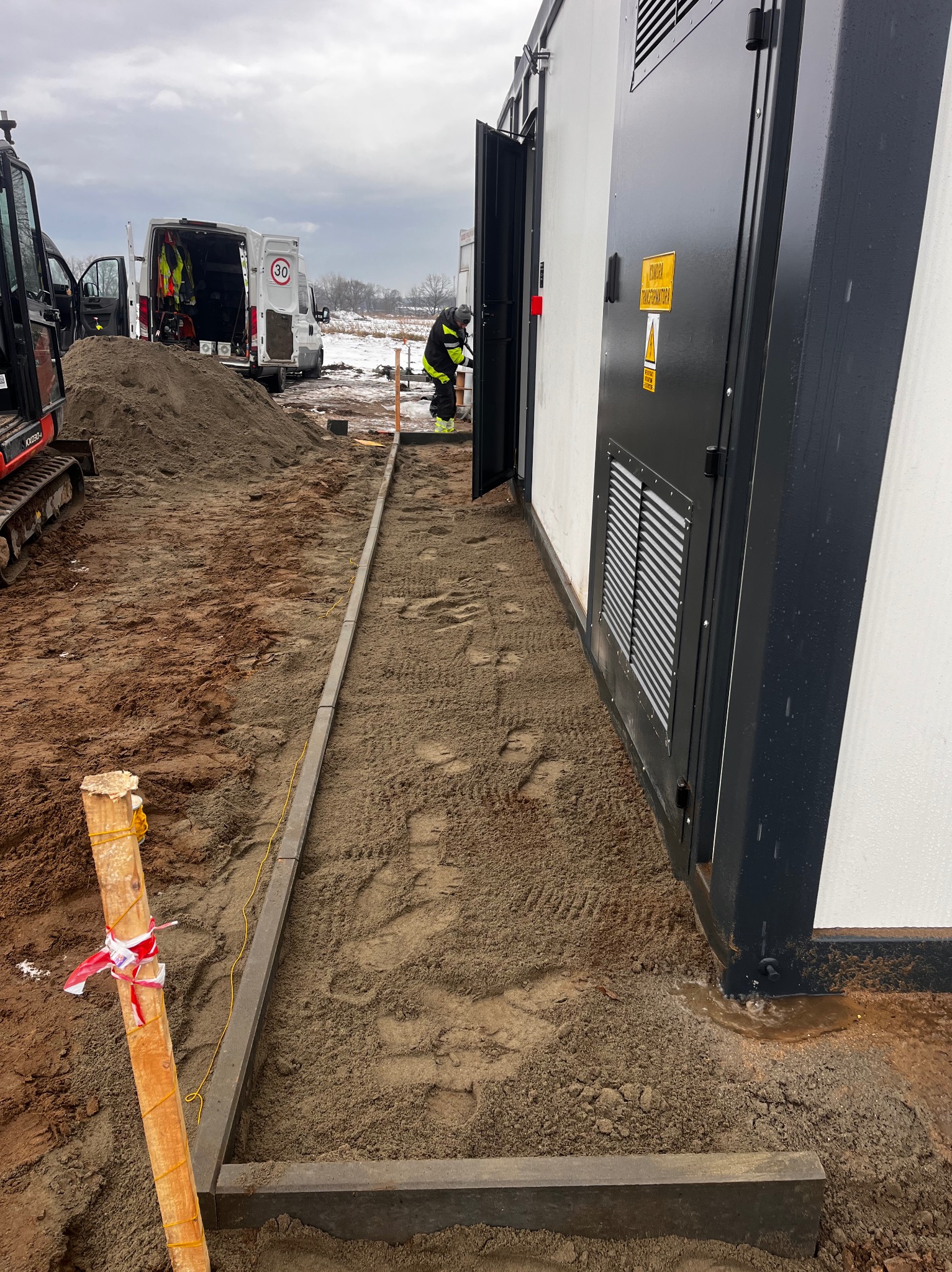 Układanie obrzeży betonowych wokół transformatora. W tle widać mini koparkę i samochody dostawcze. Prace ziemne w toku, teren przygotowany pod dalsze prace.