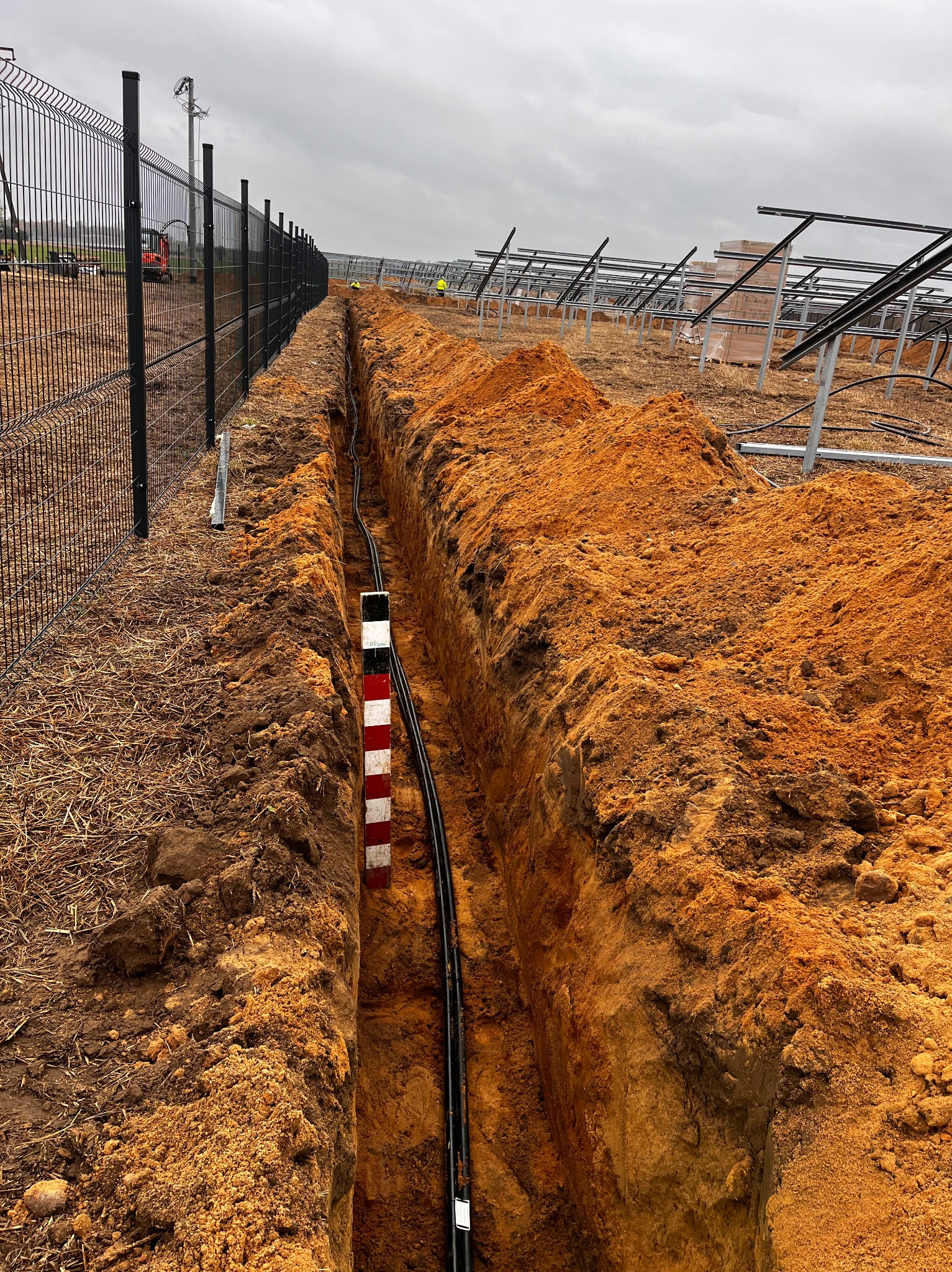 Długi wykop w ziemi z czarnymi kablami ułożonymi wzdłuż. W tle konstrukcja pod panele słoneczne i ogrodzenie z siatki. Widoczny znacznik głębokości wykopu.