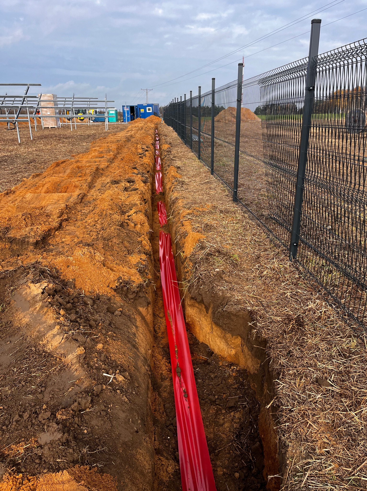 Wykop z ułożonymi czerwonymi rurami w ziemi, obok ogrodzenie z siatki. W tle widoczna konstrukcja paneli słonecznych i kontenery. Prace ziemne w toku.