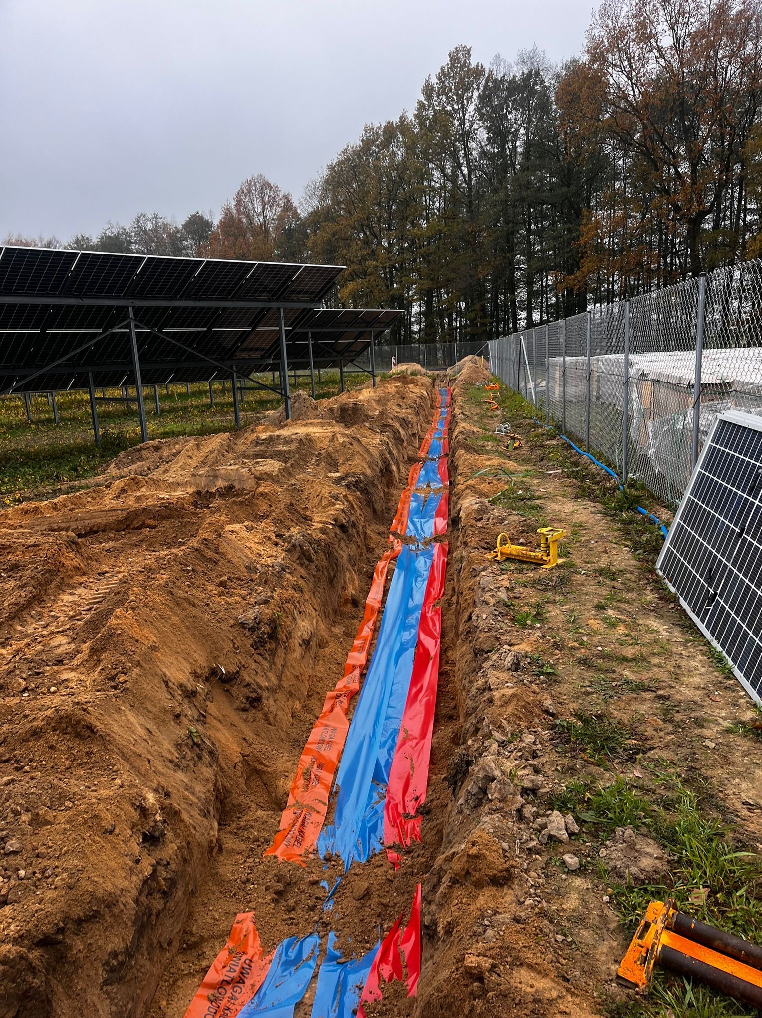 Wykop pod instalacje fotowoltaiczną zabezpieczony folią w Kędzierzynie-Koźlu. Widoczne panele słoneczne i ogrodzenie. Prace ziemne w toku, instalacja pod ziemią.