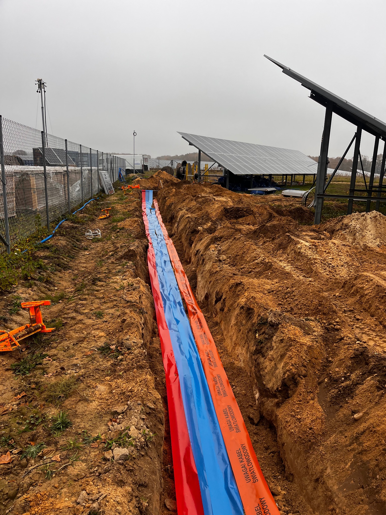 Wykop liniowy zabezpieczony folią ostrzegawczą, prawdopodobnie pod kable światłowodowe, na tle instalacji paneli słonecznych i ogrodzenia z siatki.