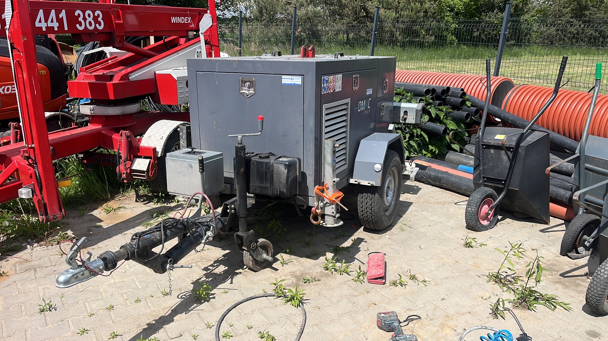 Przenośny kompresor budowlany na przyczepie, obok czerwony podnośnik i rury drenarskie. Widoczne narzędzia i wózek transportowy. Sprzęt na placu budowy.