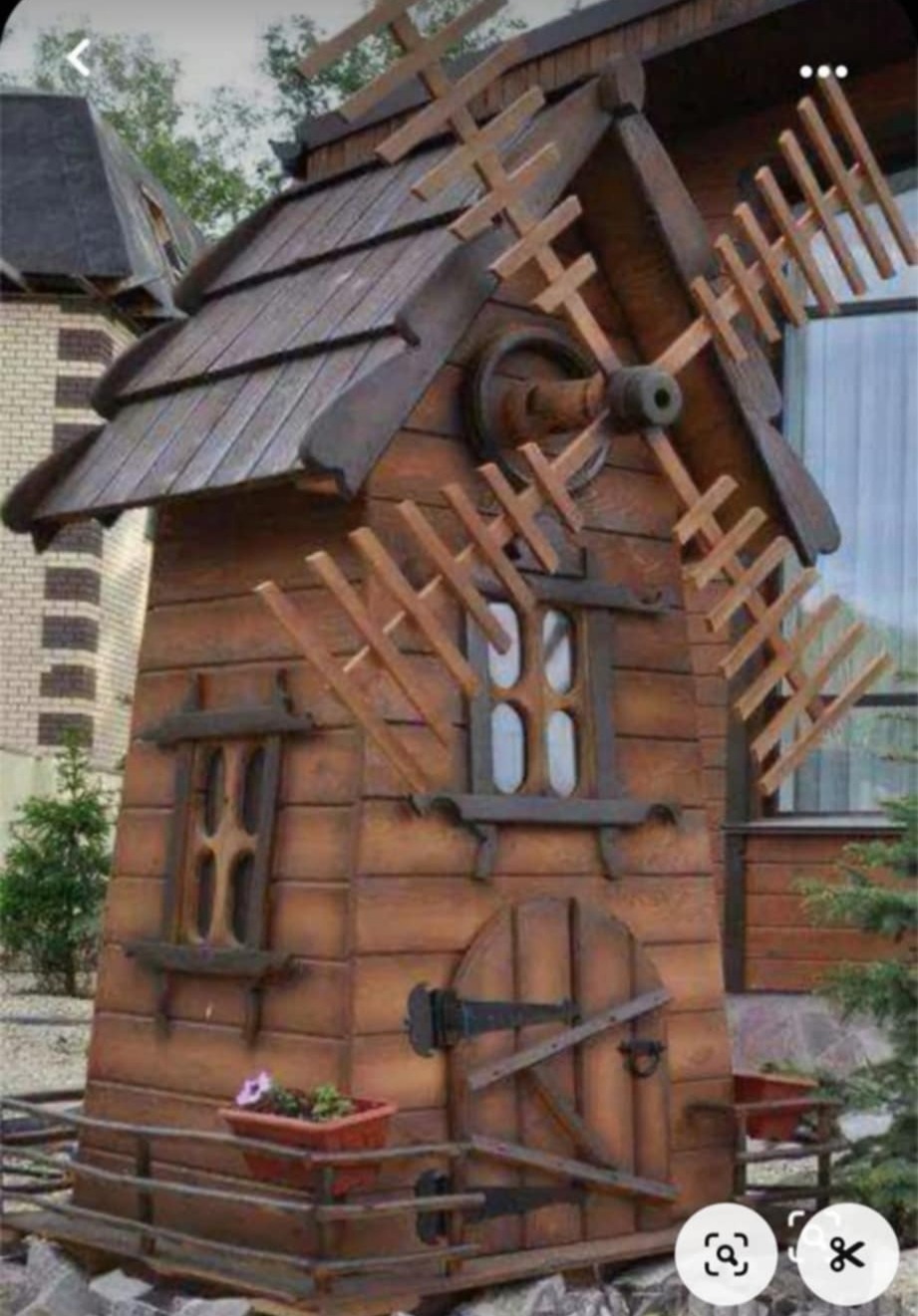 Drewniana konstrukcja w kształcie wiatraka z oknami i drzwiami, umieszczona w ogrodzie. Dekoracyjny element architektury ogrodowej, wykonany z brązowego drewna.