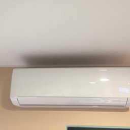 AS - CLIMATECH Arkadiusz Sosna - Biała klimatyzacja split na beżowej ścianie pod białym sufitem. Widoczne okno w dolnej części kadru. Oświetlenie dzienne.