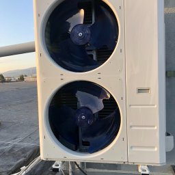 AS - CLIMATECH Arkadiusz Sosna - Biała, dwu-wentylatorowa jednostka zewnętrzna klimatyzacji Kaisai zamontowana na dachu budynku, z widocznymi przewodami. W tle krajobraz miejski o zmierzchu.